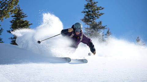  A skier on high performance skis cuts through powder, spraying snow, under a clear blue sky.