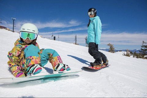 A little girl in multi-colored snow clothes rests as she makes her way down the mountain on a rented snowboard. 